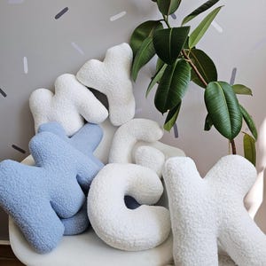 May include: A set of plush letters spelling out "MY CK" in white and light blue. The letters are arranged on a white chair with a fluffy white rug in the foreground. A green plant is in the background.