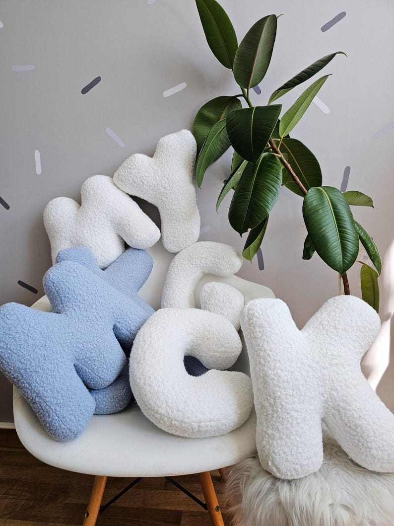 May include: A set of plush letters spelling out "MY CK" in white and light blue. The letters are arranged on a white chair with a fluffy white rug in the foreground. A green plant is in the background.