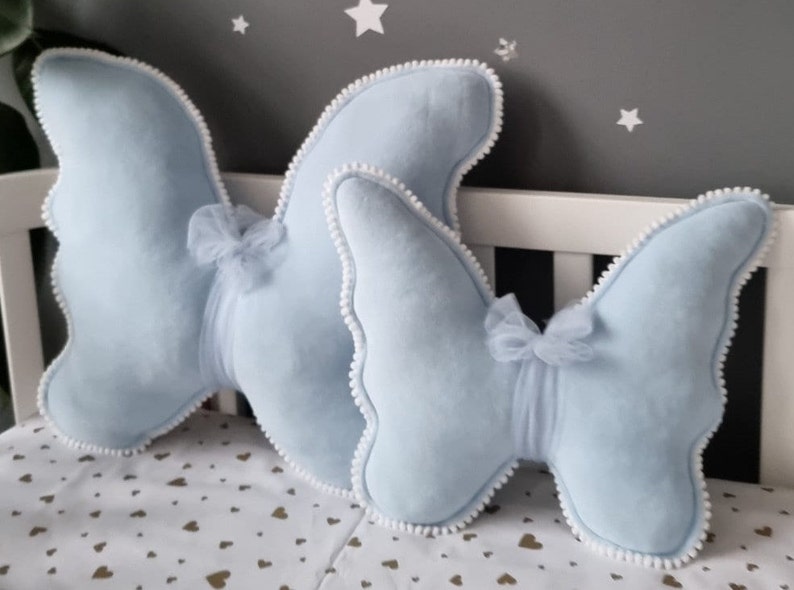 May include: Two light blue butterfly-shaped pillows with white pom-pom trim and a tulle bow in the center of each pillow.