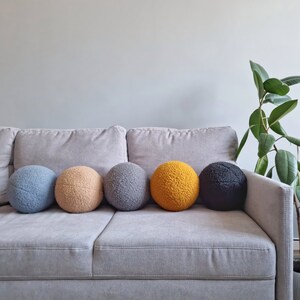 May include: Five round, fluffy pillows in different colors are arranged on a gray couch. The pillows are blue, beige, gray, yellow, and black.