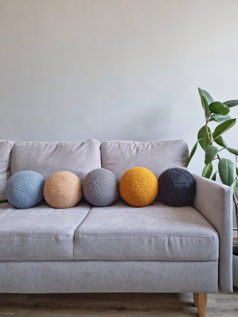 May include: Five round, fluffy pillows in different colors are arranged on a gray couch. The pillows are blue, beige, gray, yellow, and black.