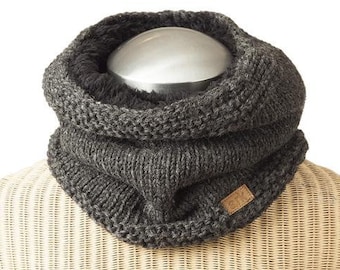 May include: A dark gray knitted cowl scarf with a soft, plush interior. The scarf is displayed on a mannequin and features a small, rectangular leather tag. The knitted design is visible, showcasing the texture and warmth of the accessory.