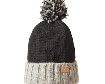 May include: A black and gray knit beanie with a large pom-pom. The hat features a ribbed gray brim and a black upper section. A small brown leather tag with the word "ark" is sewn on the brim.