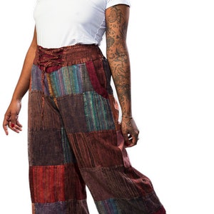 May include: A woman wearing a white t-shirt and patchwork cropped trousers with a lace-up detail at the waist. She is wearing sandals and has tattoos on her arms.