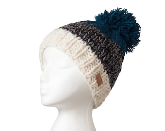 May include: A knitted beanie hat with a teal pom-pom. The hat features a cream, gray, and black striped design. A small brown tag is sewn on the side. The hat is displayed on a white mannequin head.