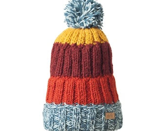 May include: A colorful knit beanie with a pom-pom. The hat features horizontal stripes in shades of blue, orange, burgundy, and yellow. A small, rectangular tag with the word "ark" is sewn on the brim.