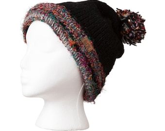 May include: Black knit beanie with a colorful, textured brim and a matching pom-pom. The brim features a mix of red, green, and purple yarns. A small brown leather tag with the word "ark" is sewn on the side. The hat is displayed on a white mannequin head.