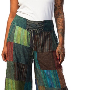 May include: A woman wearing a white t-shirt and patchwork trousers with a lace-up waistband. The trousers are made of different coloured fabrics, including green, brown, blue, and red.