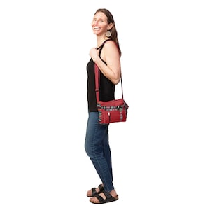 May include: A woman wearing a black tank top and blue jeans is smiling and holding a red and multi-coloured crossbody bag.