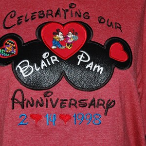 Puede incluir: Camiseta roja con orejas de Mickey Mouse bordadas en negro y un corazón con Mickey y Minnie Mouse dentro. El texto "Celebrating Our Blair Pam Anniversary 2 14 1998" está bordado en negro y azul.