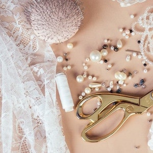 May include: A flat lay of white lace fabric, a spool of white thread, gold scissors, and pearl beads on a beige surface.