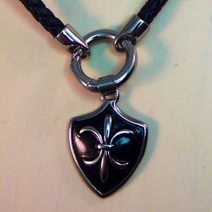 May include: A black leather necklace with a silver-colored metal pendant. The pendant is shield-shaped with a fleur-de-lis design. The necklace has a braided leather cord and a large metal ring.