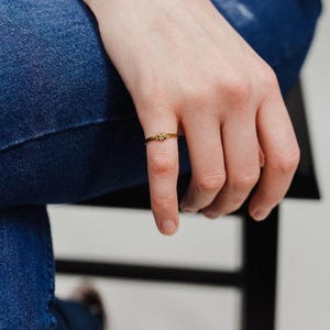 A close-up view of a person's hand wearing a gold Double Knot Ring resting on a blue denim chair.