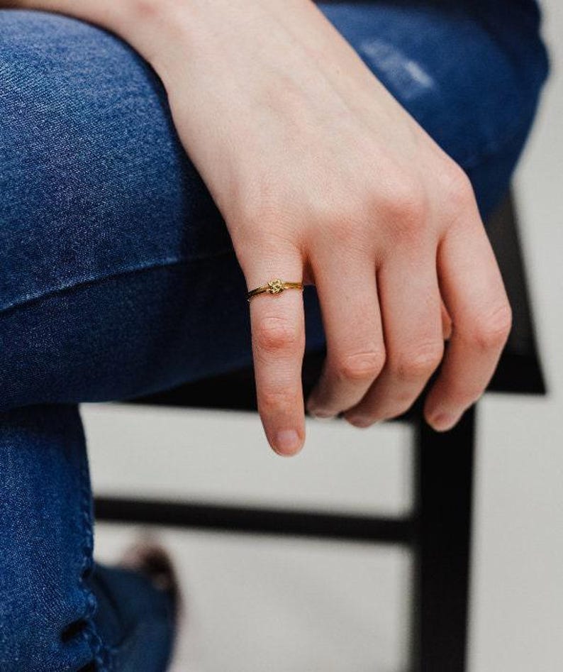 A close-up view of a person's hand wearing a gold Double Knot Ring resting on a blue denim chair.