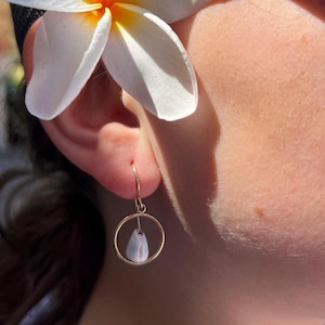 May include: Gold hoop earrings with a teardrop-shaped, light blue pendant. A white plumeria flower with a yellow center rests near the ear. The earrings are simple and elegant, suitable for everyday wear.