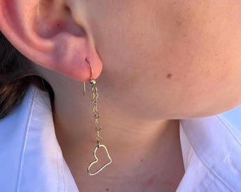 May include: A gold-coloured drop earring featuring a heart-shaped pendant. The earring has a small hook for the ear. The pendant is a delicate outline of a heart, suspended from a chain of tiny hearts.