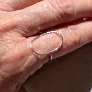 May include: A silver ring with an open, oval design. The ring is simple and elegant, with a thin band and a textured surface on the oval. The ring is shown on a hand.