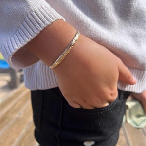 May include: A gold-colored bangle bracelet with an engraved floral design. The bracelet is worn on a wrist, and the person is wearing a white sweater and black pants. The bracelet is approximately 2 inches in diameter.