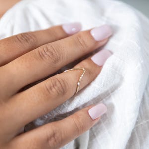 May include: A gold-toned, delicate, V-shaped ring worn on a finger with light pink nail polish.