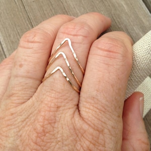 Puede incluir: Un conjunto de tres anillos dorados en forma de chevrón. Los anillos están apilados en un dedo, mostrando su textura martillada y diseño geométrico. Los anillos son de un tono dorado cálido, reflejando la luz.