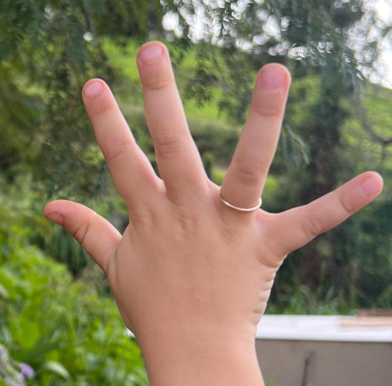 May include: A close-up of a hand wearing a thin silver ring on the ring finger.