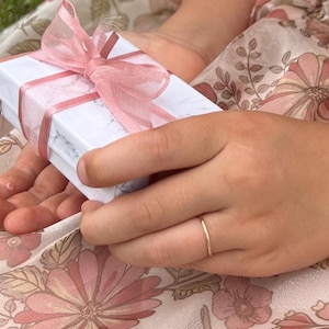 Peut inclure: Une petite boîte cadeau blanche avec un motif marbré est attachée avec un ruban rose. Une fine bague en or est visible sur un doigt. L'arrière-plan présente un motif floral rose et crème.