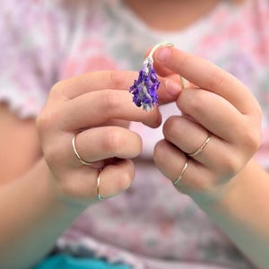 May include: A person's hands wearing two thin gold rings, holding a dried purple flower.