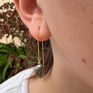 Puede incluir: Un pendiente de cadena dorada con una perla barroca gris oscura. El pendiente es de estilo enhebrador, con la cadena que atraviesa la oreja y la perla colgando debajo. El pendiente se muestra en la oreja de una persona.