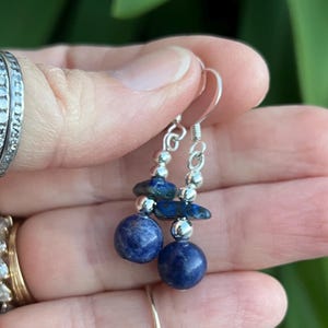 Puede incluir: Pendientes colgantes con cuentas esféricas azul oscuro y piedras azules pequeñas de forma irregular. Los pendientes tienen detalles metálicos plateados y ganchos para las orejas. El diseño es sencillo y elegante.