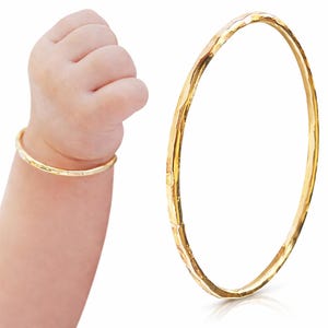 Puede incluir: Un brazalete dorado con textura martillada. El brazalete se muestra en la muñeca de un bebé, y un brazalete separado se muestra sobre un fondo blanco. El brazalete es una joya sencilla y elegante.