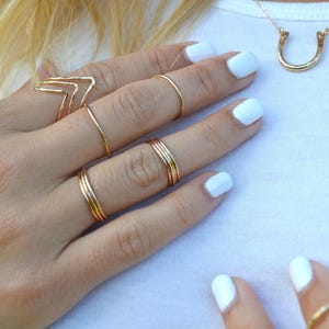 Puede incluir: Una mano muestra varios anillos dorados de diferentes diseños, incluyendo un galón y bandas finas. También se ve un collar con un colgante de herradura dorada. Las uñas están pintadas de blanco. La joyería se presenta sobre una camiseta blanca.