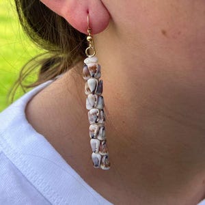 May include: A long, dangling earring featuring a vertical column of small, natural, brown and white speckled seashells strung together. The earring is attached to a thin, gold-toned metal hook that passes through the earlobe.
