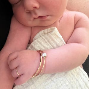 Puede incluir: Una pulsera dorada con un acento de perla y un anillo dorado en la mano de un bebé. La pulsera tiene una textura martillada. El bebé está envuelto en una tela de muselina blanca.