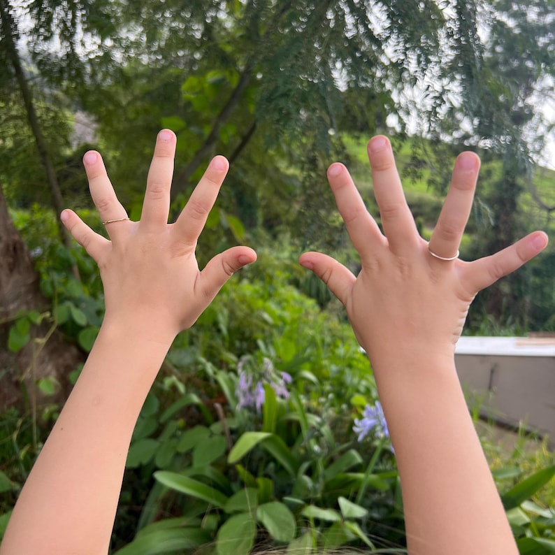 May include: Two hands with fingers spread out, reaching towards the sky. The hands are wearing thin gold rings on the ring fingers.