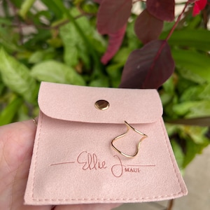 Puede incluir: Una bolsa de tela rosa claro similar a la gamuza con un cierre de bot&oacute;n a presi&oacute;n dorado. La bolsa presenta el texto en relieve Ellie J Maui. Un anillo de alambre dorado fino con un dise&ntilde;o ondulado y abierto descansa en la parte frontal.