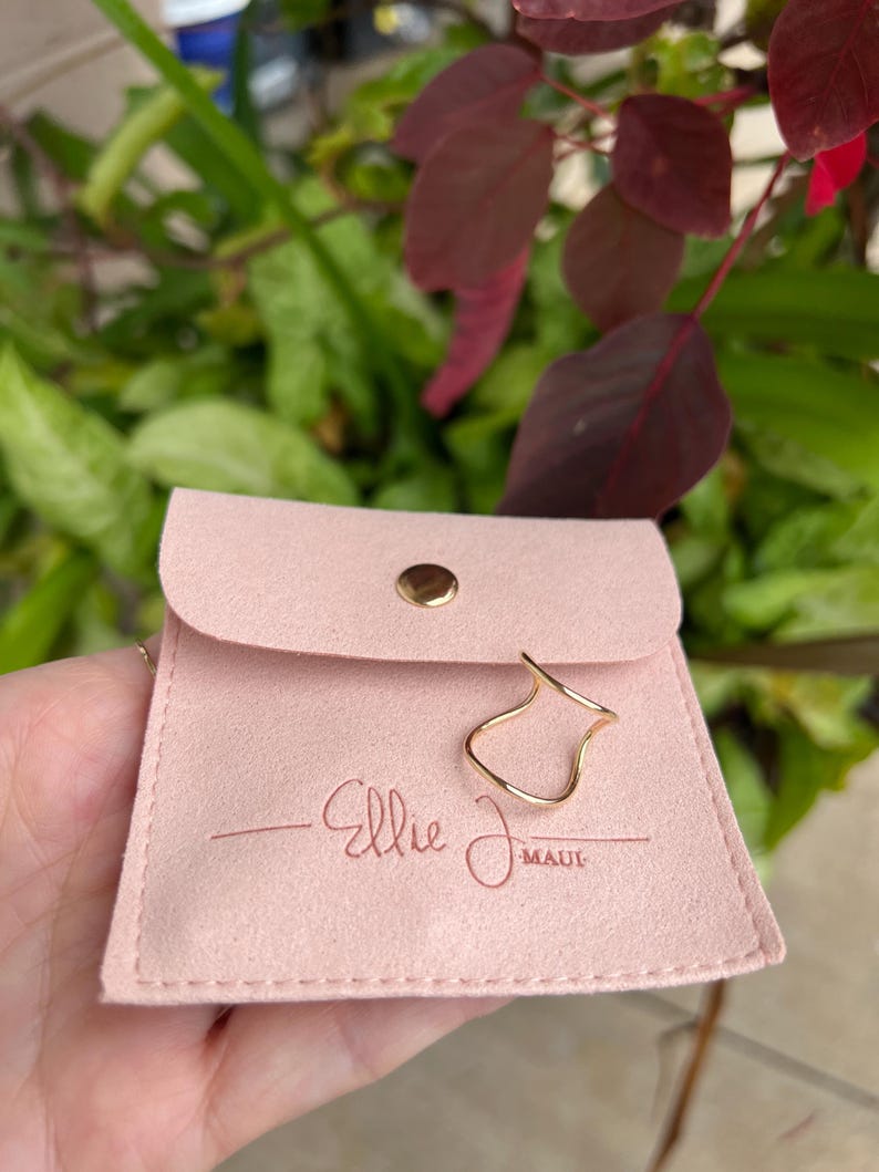 Puede incluir: Una bolsa de tela rosa claro similar a la gamuza con un cierre de bot&oacute;n a presi&oacute;n dorado. La bolsa presenta el texto en relieve Ellie J Maui. Un anillo de alambre dorado fino con un dise&ntilde;o ondulado y abierto descansa en la parte frontal.