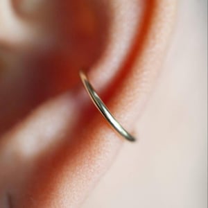 May include: A close-up of a gold-colored ear cuff. The thin, curved jewelry piece is positioned on the ear, with the skin in the background. The cuff has a smooth, polished surface and a simple, elegant design.