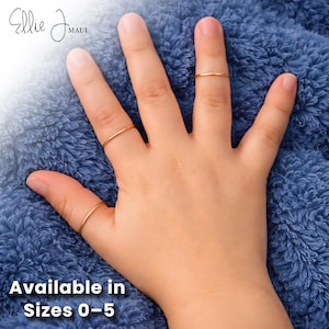 May include: A hand wearing three thin, gold-colored rings. The rings are simple bands. The text "Available in Sizes 0-5" is at the bottom of the image. The brand name "Ellie J Maui" is in the top left corner.
