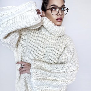 May include: A white oversized knitted sweater with a turtleneck and long sleeves. The sweater has a chunky knit texture and a relaxed fit.
