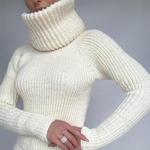 May include: Cream-colored turtleneck sweater with a ribbed knit design. The long-sleeved sweater has a high, folded collar and a fitted silhouette. The knitwear appears to be made of a thick, warm yarn, suitable for cooler weather.