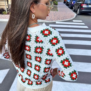 May include: White crochet sweater with red and green floral pattern. The sweater has long sleeves and a crew neckline.