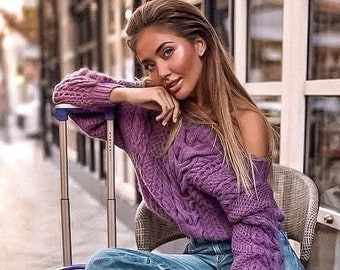Könnte beinhalten: Eine Frau in einem lila Zopfstrickpullover und blauen Jeans sitzt auf einem Stuhl mit einem lila Koffer. Der Pullover hat schulterfreie Ärmel. Der Koffer hat einen Teleskopgriff. Die Frau trägt schwarze Stiefel.