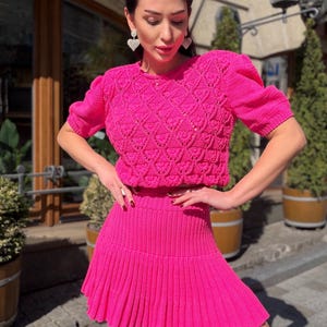 May include: A bright pink, two-piece knit outfit. The top is a short-sleeved, cropped sweater with a textured pattern. The matching skirt is pleated. The outfit is a vibrant, eye-catching ensemble.