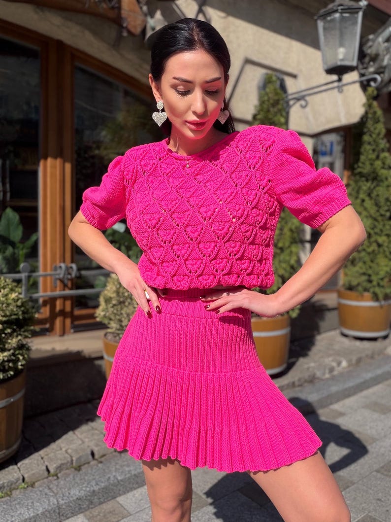 May include: A bright pink, two-piece knit outfit. The top is a short-sleeved, cropped sweater with a textured pattern. The matching skirt is pleated. The outfit is a vibrant, eye-catching ensemble.