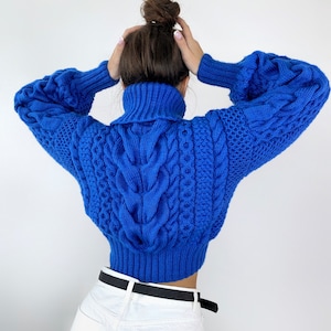 Könnte beinhalten: Ein leuchtend königsblauer Zopfmuster-Pullover mit hohem Rollkragen, langen Ärmeln mit gerippten Bündchen und kurzem Saum. Das Strickmuster zeigt komplexe vertikale Zopfstrukturen und Seitenteile mit einem Wabenmuster-Design.