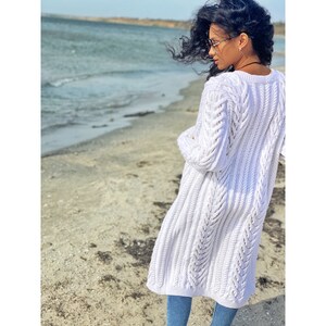 May include: A long, white cable-knit cardigan with a textured design. The cardigan is worn over blue jeans. The image is taken on a beach with the ocean in the background.