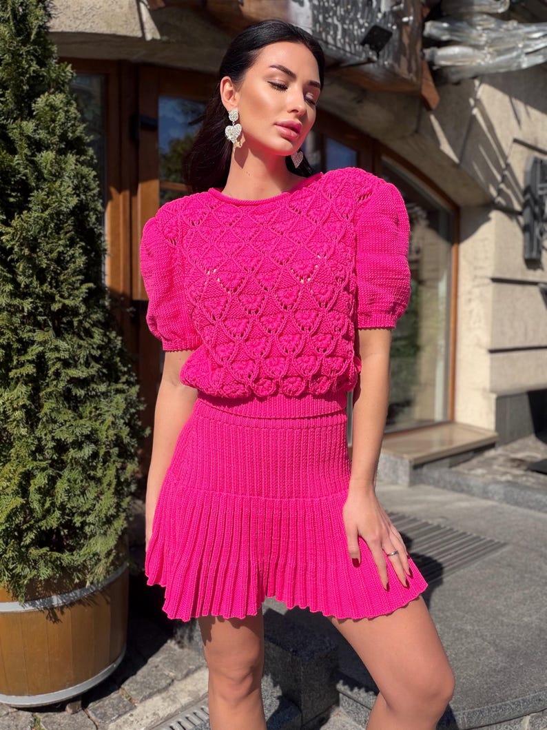 May include: A bright pink, two-piece knit set. The top has short, puffed sleeves and a textured pattern. The skirt is pleated. The outfit is worn with heart-shaped earrings.