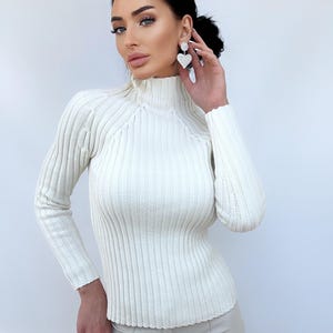 May include: A white ribbed turtleneck sweater with long sleeves. The sweater is fitted and has a high neckline.