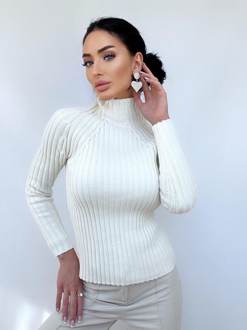 May include: A white ribbed turtleneck sweater with long sleeves. The sweater is fitted and has a high neckline.