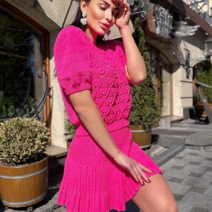 May include: A bright pink knitted two-piece outfit. The top has short sleeves and a detailed openwork pattern on the bodice. The matching skirt is a mini-skirt with a pleated hem. The outfit is made of a textured knit fabric.